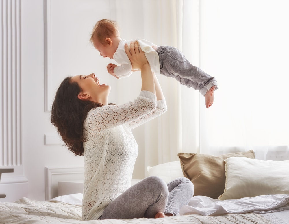 Woman holds her baby up in the air and smiles, feeling happy with her body after a mommy makeover